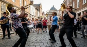 street band with brass instruments performing to a joyful, dancing crowd on a historic street during the Durham Brass Festival