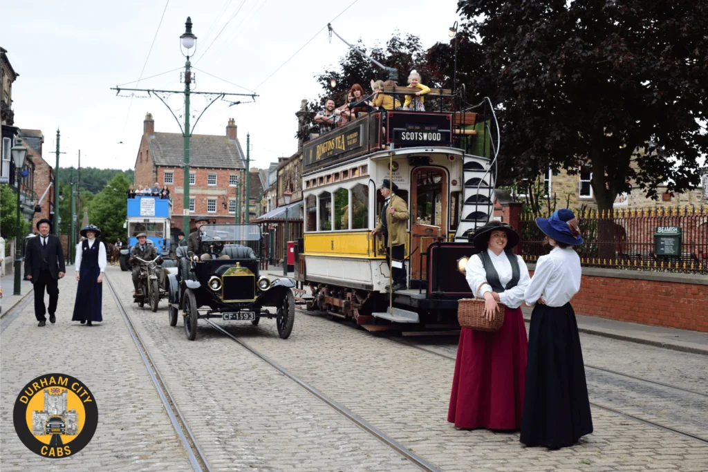 Visitors in period costumes at Beamish Museum with a vintage tram, classic car, and historic buildings on a cobbled street.