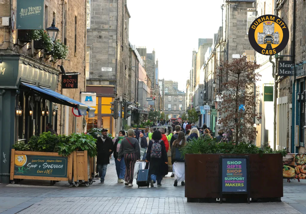 A vibrant, narrow street in Durham city centre with many people walking, historic stone buildings, shops like "Booty Cow" and "Meze Mezze," and outdoor planters. The Durham City Cabs logo is visible in the top right.