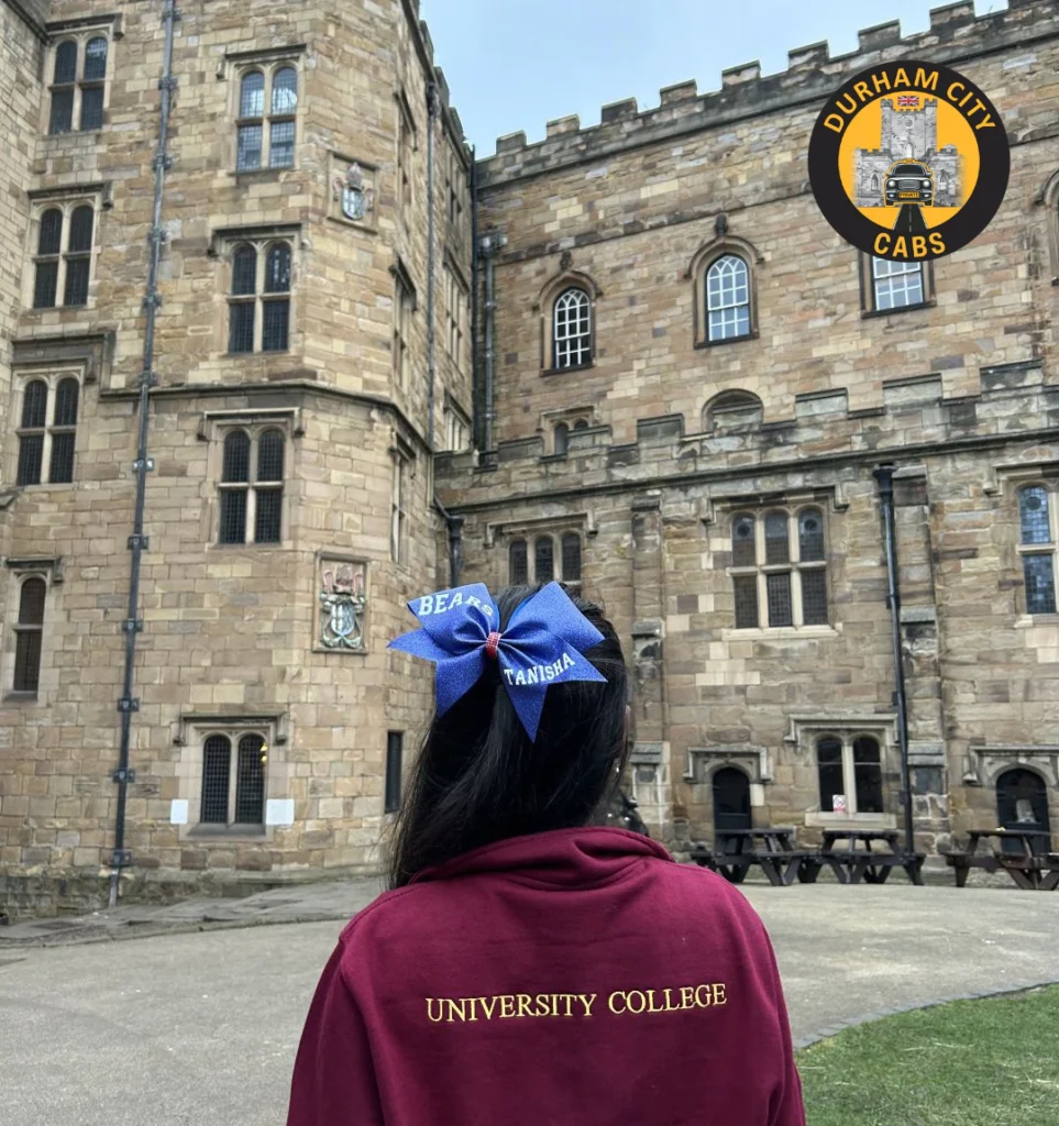 Back view of a student wearing a "University College" hoodie and a blue bow, standing in front of the historic Durham Castle (University College) at Palace Green.