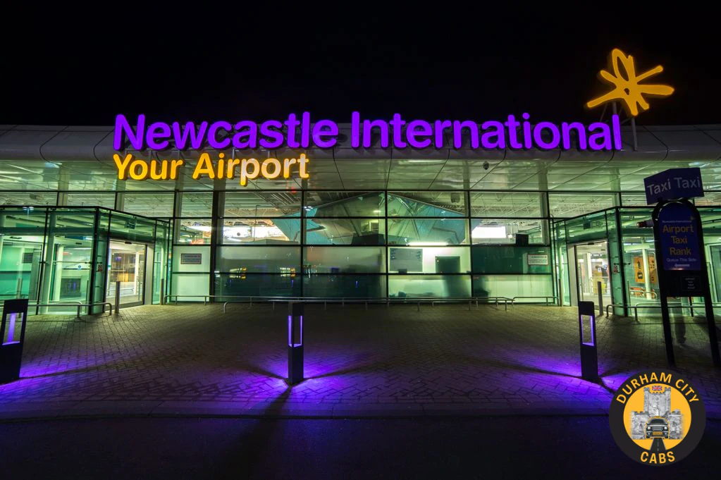 Illuminated entrance of Newcastle International Airport at night, showing "Newcastle International Your Airport" sign in purple and yellow lights, with the Durham City Cabs logo in the corner.