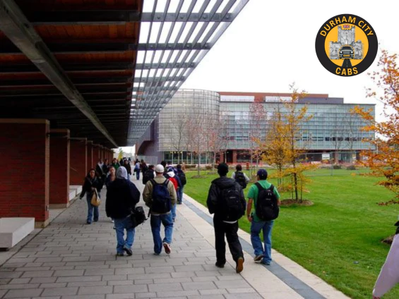 Group of students walking along a modern pathway with green lawns and contemporary university buildings in the background, representing Durham's Hill colleges.